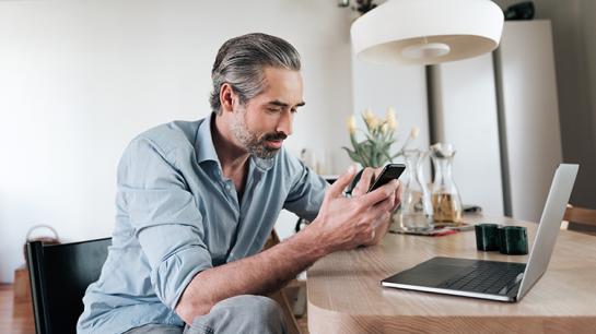 Ein Mann sitzt vor einem Laptop und blickt auf sein Smartphone.